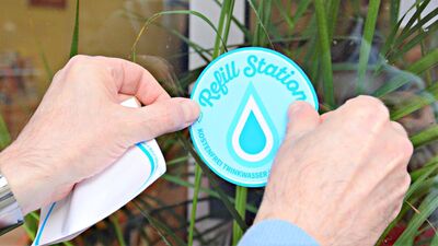 Zwei Hände kleben einen Sticker mit dem „Refill Station“-Logo an ein Fenster.